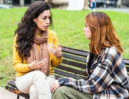 Two women sit on a park bench. One gestures as the other leans in to listen.