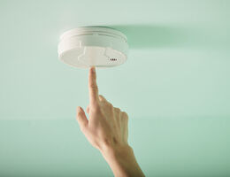 A hand reaches up to push the test button on a smoke alarm.
