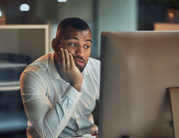A man in a dark office rests his head on his hand as he looks at his computer screen.