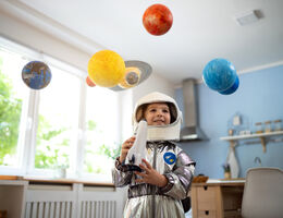 A child in an astronaut costume holds a rocket under a solar system mobile.